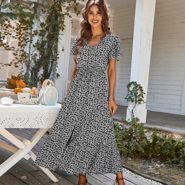 A woman standing outdoors wearing a floral print long dress with a V-neck and short sleeves. The dress is black with a pattern of floral designs.
